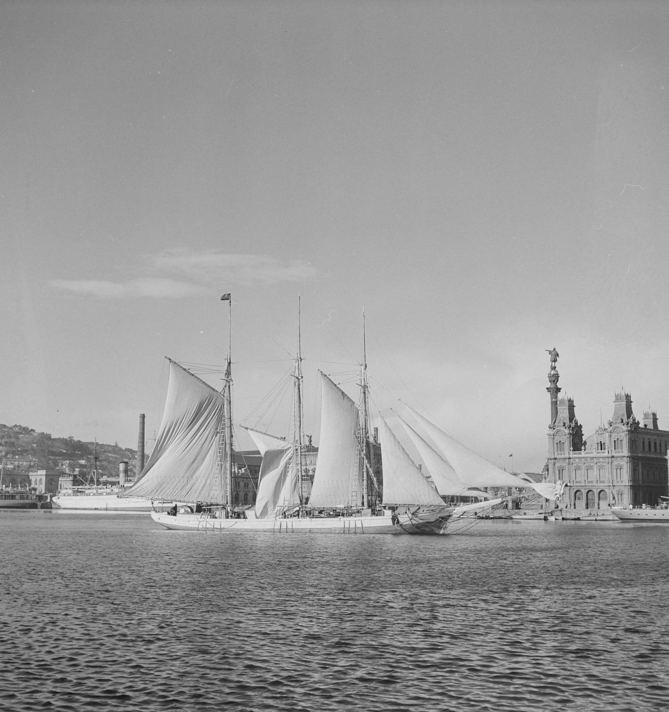 Velero de tres palos con velas desplegadas en el puerto de Barcelona, con el Port Vell y el Mirador de Colón al fondo. Joaquín Tusquets de Cabirol, c. 1950.